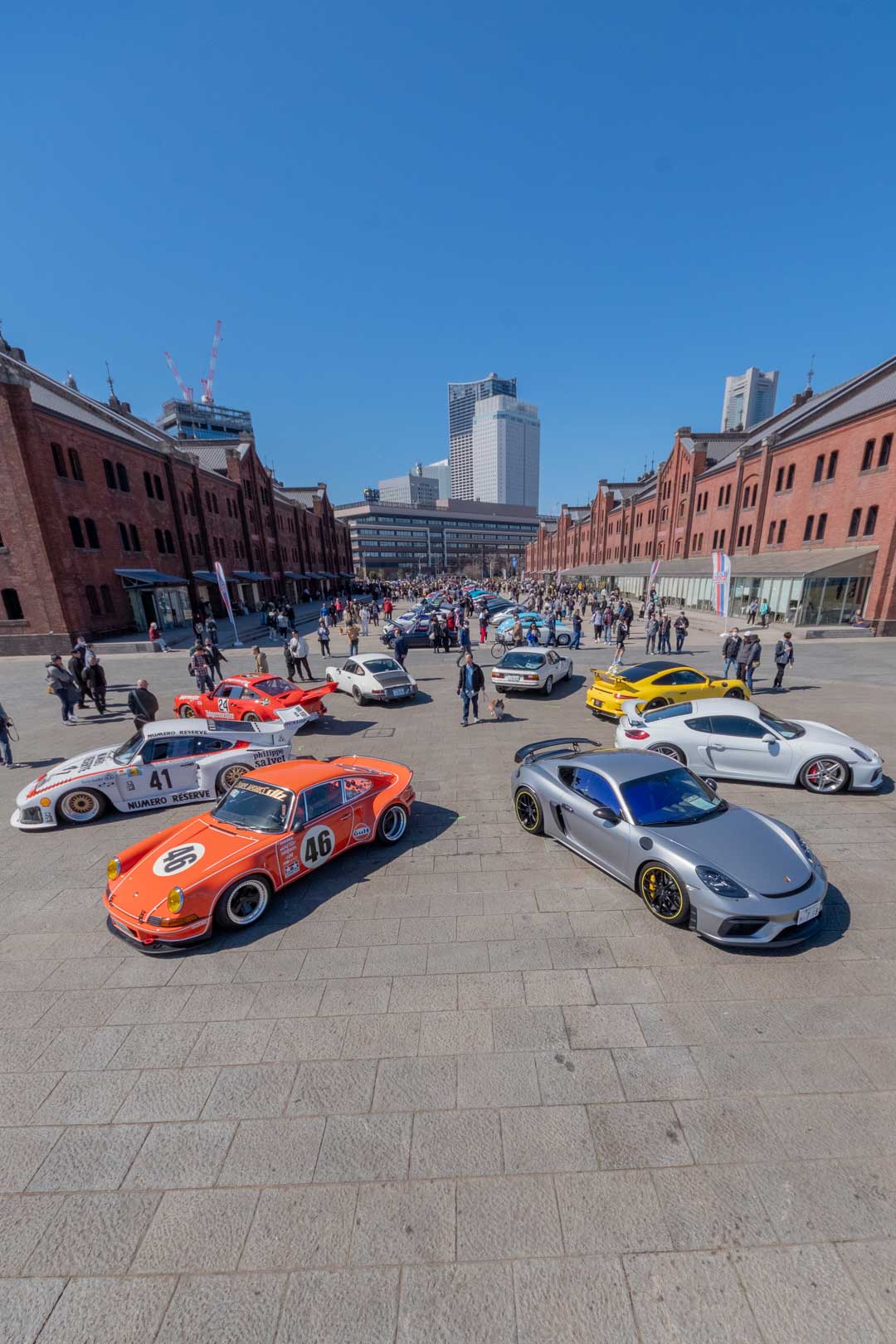 2026 年 EXCITING PORSCHE（横浜 赤レンガ倉庫）特別展示エリアの配置例 1。赤レンガ倉庫の間の広場に、レーシングカラーの 911 やケイマンが向かい合わせに並ぶ様子