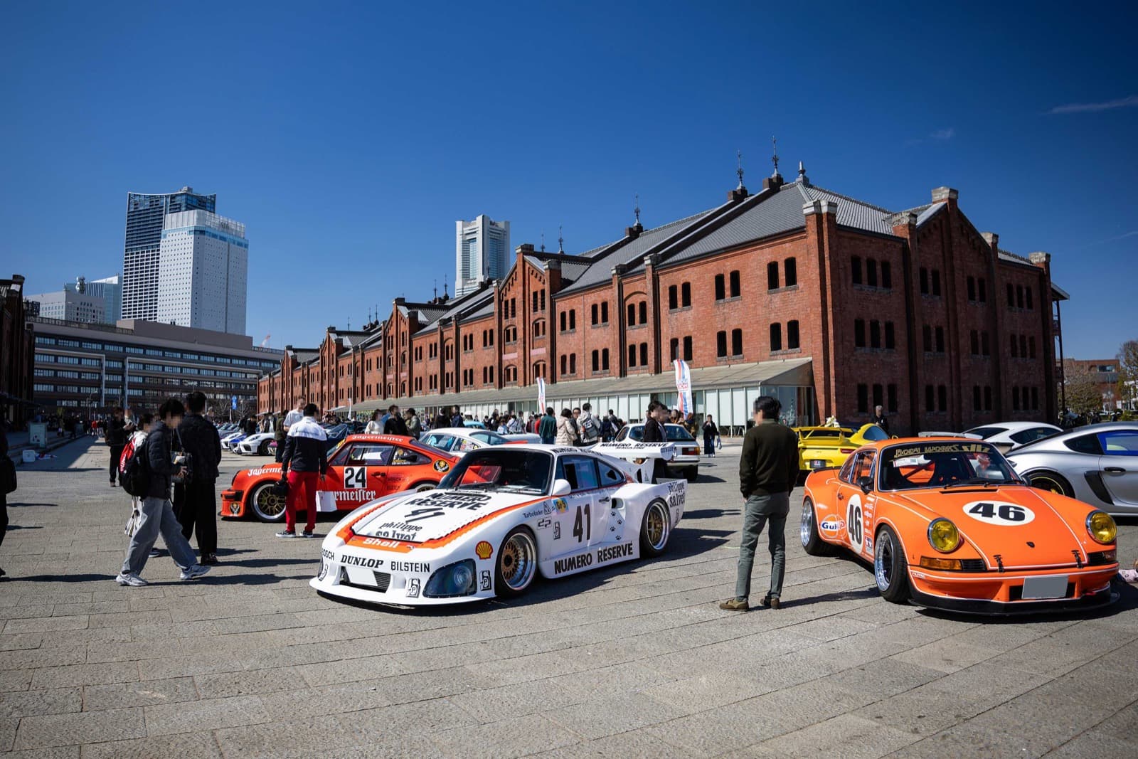 2026 年 EXCITING PORSCHE（横浜 赤レンガ倉庫）特別展示エリアの配置例 4。赤レンガ倉庫を背に、白いレース仕様のポルシェとオレンジの 911 が並ぶ様子
